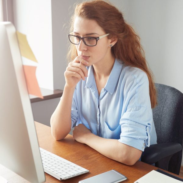 Confident female freelancer sitting in front of the computer with serious and thoughtful expression. Young businesswoman in blue shirt and glasses using PC for work sitting in the office